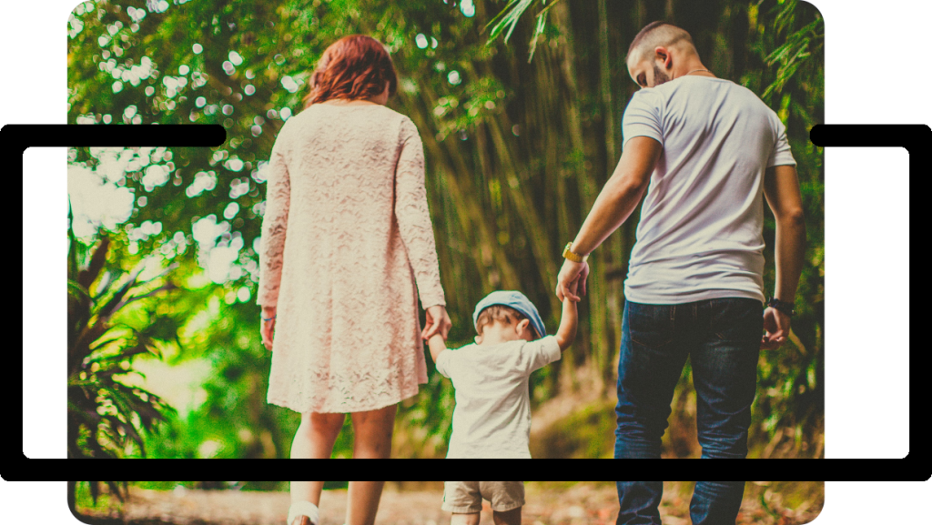 Mother and father holding hands with their son, walking down a wooded path.